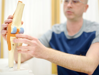 Orthopedist shows the patient an anatomical model of knee joint during appointment in modern...