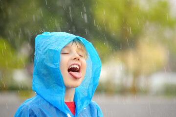 Portrait of a happy child in the pouring rain. A joyful boy in a blue raincoat catches water drops...