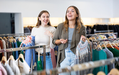 Two young cheerful woman shopping together, searching casual clothes for cold days in showroom and...