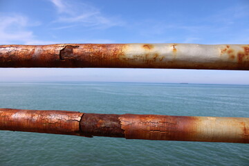 A rusty fence with a blue sea in the background.Rusty iron railing, beautiful sea and sky landscape view between rusty railing gap.