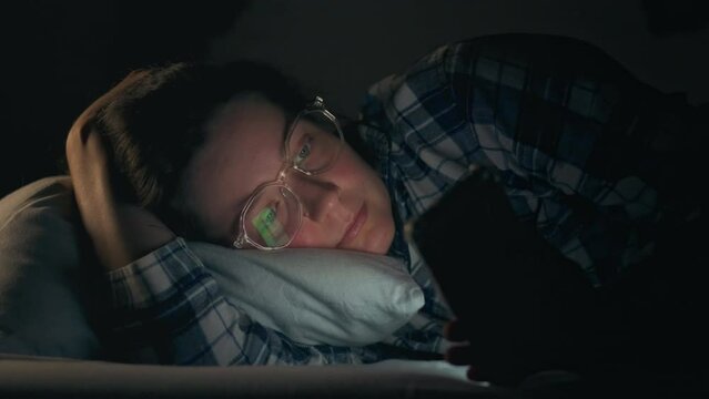 A young girl of European appearance lying in bed at night looks at the bright screen of a smartphone. Woman can't take her eyes off while consuming social media information, smartphone addiction