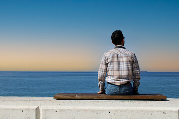 Young sad depressed man sit alone on bench, looking at distant sea,seascape horizon. Time to go, say goodbye. Miss someone. hope to go far away
