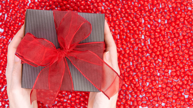 Female's hands holding Christmas gift box decorated with red ribbon bow on red candy background. Christmas and New Year banner. Christmas shopping, Boxing day, pepermint candies.