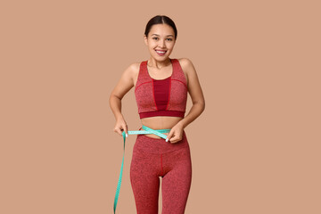Young woman in sportswear measuring her waist on beige background. Weight loss concept