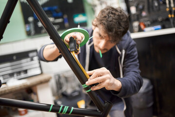Man taking measurements with a tape measure, process of preparing a bicycle frame with masking tape for a custom painting design in a bike workshop. Selective focus composition. Real people working.