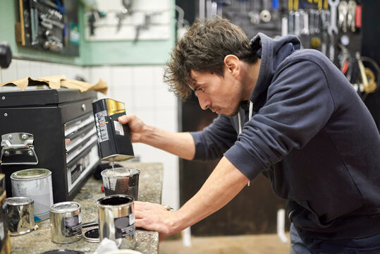 Young hispanic man pouring thinner into a cup of black paint on a workshop countertop. Real people at work.