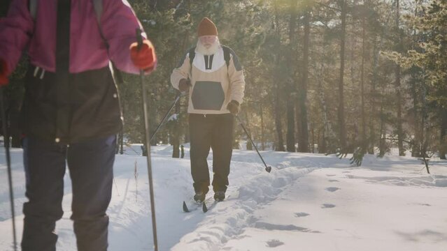Full lockdown shot with lens flare of elderly Caucasian wife in woolly hat, jacket and husband with thick grey beard, in anorak, with backpack enjoying ski trip together in forest on sunny day