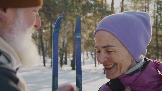 Medium close-up shot of loving senior Caucasian man zipping up jacket for smiling wife, straightening hat, chatting, while skiing together in snowy forest during winter vacation