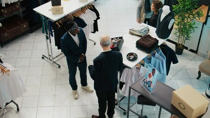 Senior designer teaching his apprentice to match ties on elegant suits, presenting the color mixing theory to his pupil. African american man trying to learn basics in fashion industry. Camera B.