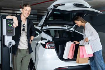Young couple travel with EV electric car to shopping center parking lot charging in downtown city...