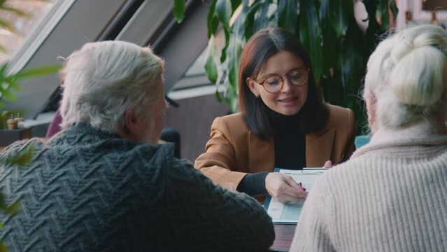 Medium over shoulder shot of female financial adviser having business meeting with senior clients in cafe, presenting insurance policy, explaining legal details, husband and wife listening, nodding