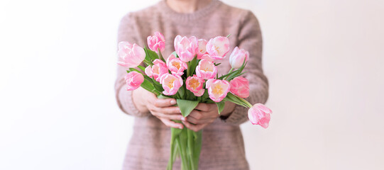 Unrecognizable woman in brown knitted sweater holds bouquet of pink tulips in her hands and extends them forward to the camera. Focus on flowers in foreground. Banner, space for text.