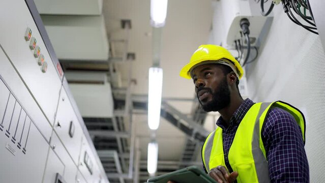 African man engineer in safety uniform working on digital tablet at factory server electric control panel. Industrial technician worker maintenance checking power system at manufacturing plant room.
