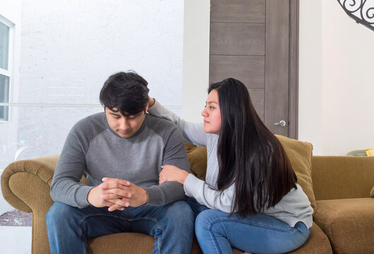 Esposa motivando a su marido tom&aacute;ndolo del brazo. Joven pareja sentada en el sof&aacute; de la sala