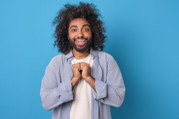 Young cheerful positive Arabian man with wide smile looks at camera clenching fists in front of chest hoping for good birthday present or cool bonus in online store stands on blue background.