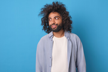 Young cheerful handsome Arabian man with curly hair and beard looks to side recommending good barbershop for creating stylish hairstyle or caring for mustache stands on blue background.