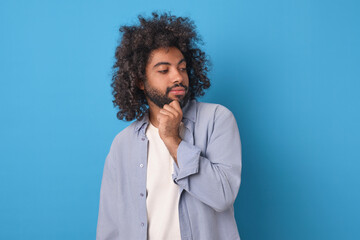 Young positive thoughtful Arabian man millennial scratching beard thinking about meaning of life or career plans and ways to ensure personal growth stands on isolated blue background.