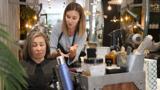 Friendly young woman hairdresser discussing haircut with aged female client sitting in chair in hairdressing salon