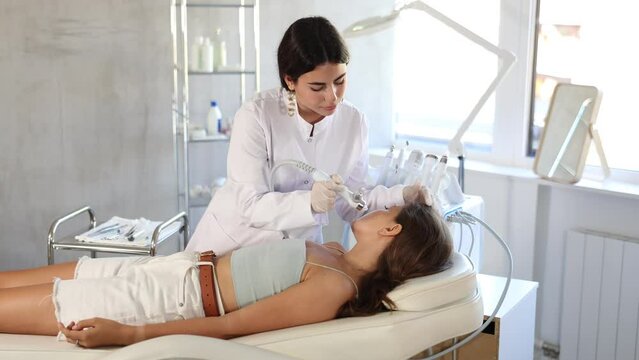 Positive young female aesthetician applying atomized nutrients with airbrush onto woman client face skin to enhance and revitalize complexion. Modern hardware cosmetology . High quality 4k footage