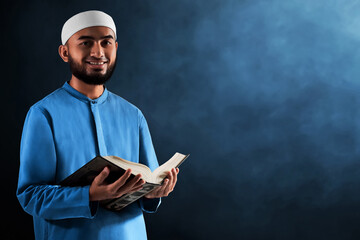 Portrait of handsome young asian muslim man with beard holding holy book quran and smiling isolated on dark blue background - 739687658