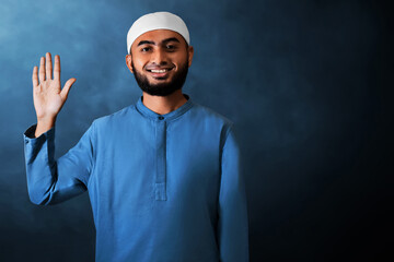 Portrait of handsome young asian muslim man with beard saying hello,  waving hand gesture and smiling isolated on dark blue studio background - 739687684