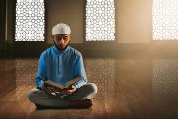 Young asian muslim man with beard reading holy book quran in the mosque window arch - 739689275