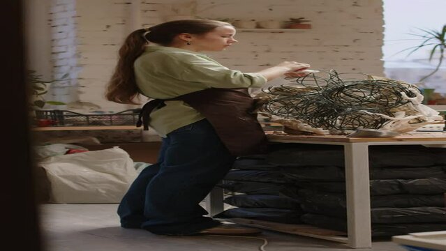 Vertical full shot of young Caucasian female sculptor in apron standing by work desk and wrapping plaster cloth around wire armature while creating piece of contemporary art in workshop