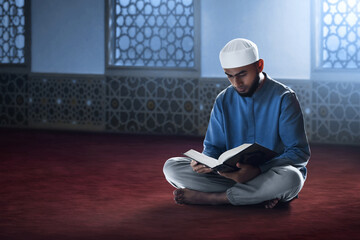 Young asian muslim man with beard reading holy book quran in the mosque window arch at night - 739690651