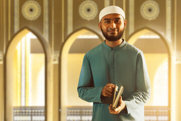 Portrait of handsome young asian muslim man with beard posing , holding holy book quran and smiling in the mosque door arch - 739690899