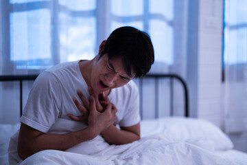 Asian anguished man clutch his chest acute pain from heart attack while sitting on a bed and wake up at night possibly immediate need help medical emergency attention in bedroom - obrazy, fototapety, plakaty