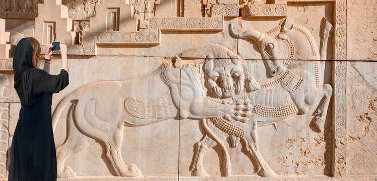 Happy Iranian girl wearing abaya taking photo - An ancient relief on the wall of the ruined city of Persepolis. A lion hunting a bull - Persepolis, Iran