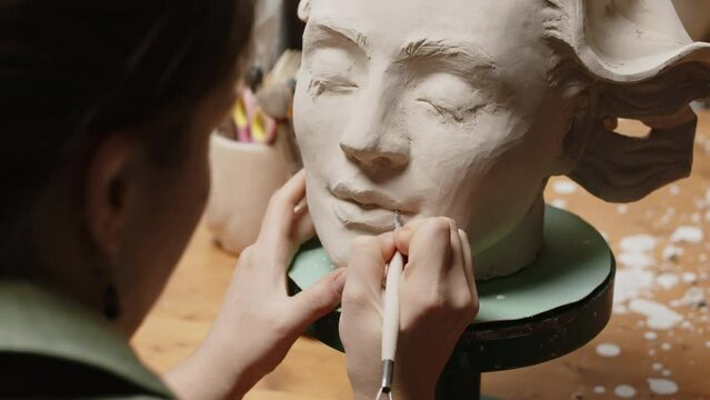 Over shoulder of female sculptor using sculpting tool while creating bust of young beautiful woman of clay in workshop