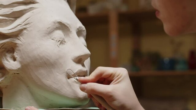 Closeup of unrecognizable female sculptor using special modeling tool while making clay woman face portrait in workshop