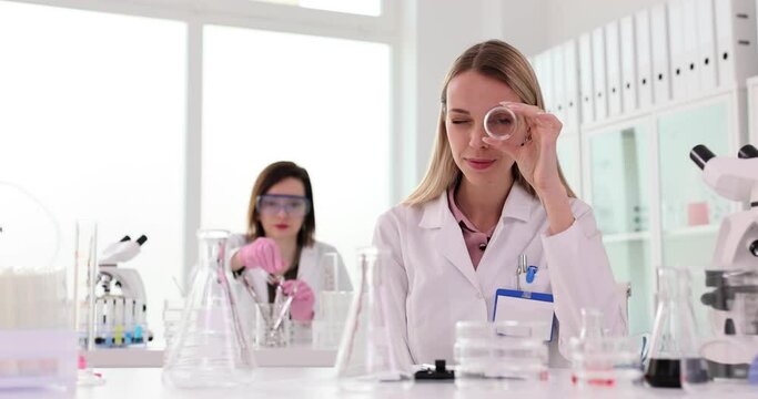 Woman looks through beaker while colleague works in lab