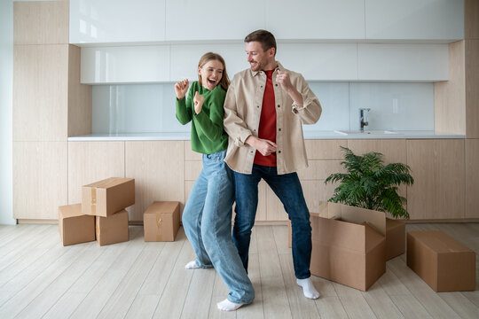 Dancing of joy and happiness, overjoy married couple have fun in spacious apartment with packed cardboard boxes. Pleased homeowners celebrating moving into own new purchased flat. Dream family home.