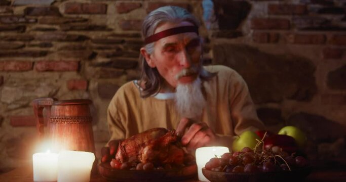 Elderly seigneur tears leg of chicken at medieval party. Grey haired aristocrat with goatee eats baked meat at dining table in tavern. Rich treat for inn guest