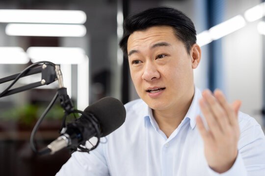 Close-up photo of an Asian young man sitting in the office at a desk in front of a microphone and having an online meeting via video call, discussing, explaining, gesturing with his hand.