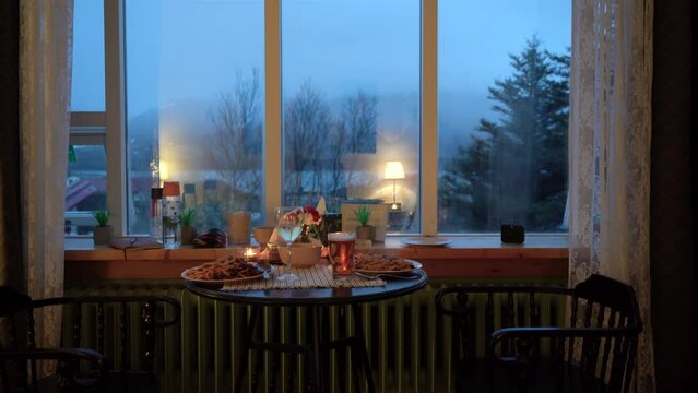 Cozy evening dinner for two by window with mood lighting, pasta and drinks on table, overcast outdoor view