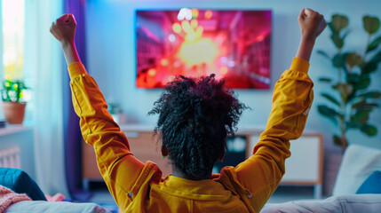 Young black African American woman enjoy a victory playing video games on TV screen raising her arms in the air on her home couch