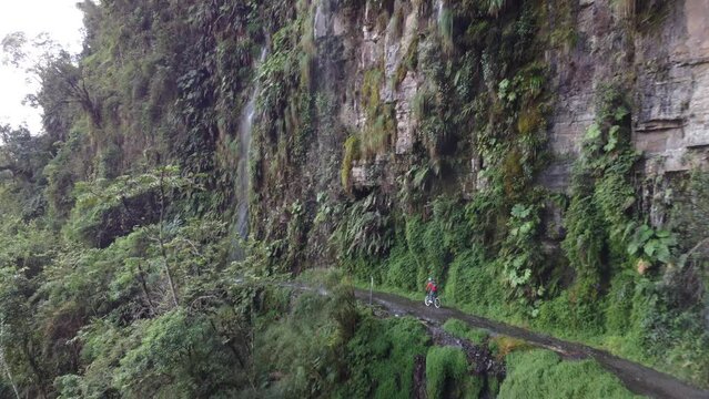 Aerial follows bicyclist where waterfall drops onto Yungas Rd, Bolivia