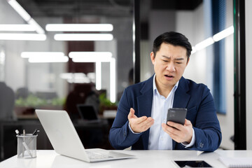 Sad disappointed asian man at workplace, dissatisfied employee in business suit with phone in...