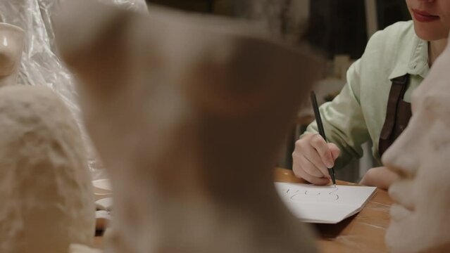 Cropped shot of unrecognizable female sculptor drawing sketch while sitting at wooden desk in sculpting workshop with many plaster head and body clay sculptures on it