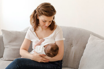 Positive young mother in casual clothes with dark hair breastfeeding newborn baby while sitting on comfortable couch in living room at home