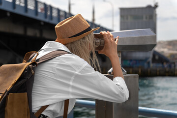 Beautiful young tourist woman in a hat with a craft backpack looks through binoculars on Galata...