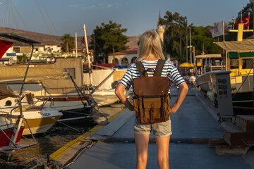 Young female tourist with a craft backpack stands on the pier near yachts and boats at sunset, back photo, Marmaris, Turkey. Concept of travel and tourism
