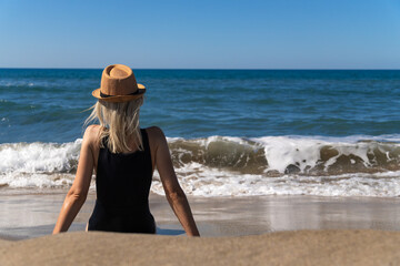 Fototapeta na wymiar Slender young woman in a black swimsuit with a hat is sitting on a sandy beach opposite the sea with waves on a sunny day, rear view. Young girl is thinking and relaxing by the sea.
