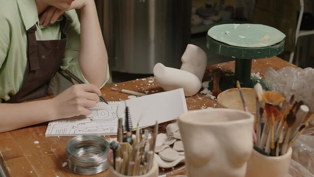 Tilt down of concentrated Caucasian female artisan drawing in sketchbook while sitting at messy wooden desk in sculpting and pottery workshopTilt down of concentrated Caucasian female artisan drawing 