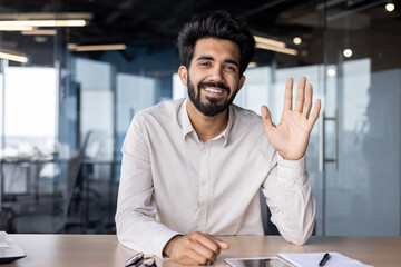 Online business meeting and training. Indian young businessman sitting at the desk in the office, waving to the camera and smiling