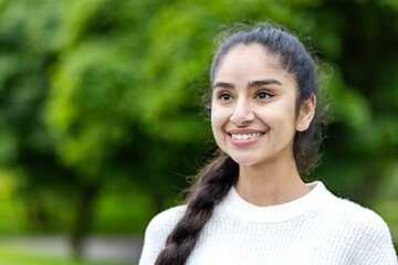 Close-up photo of a happy young Indian woman standing outside the park, smiling, dreamy and...