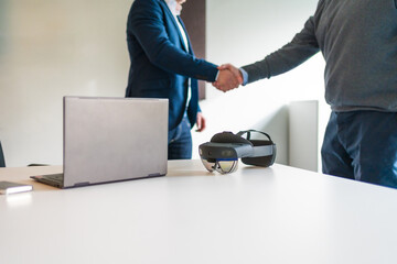 Business hand shake next to laptop and augmented reality goggles